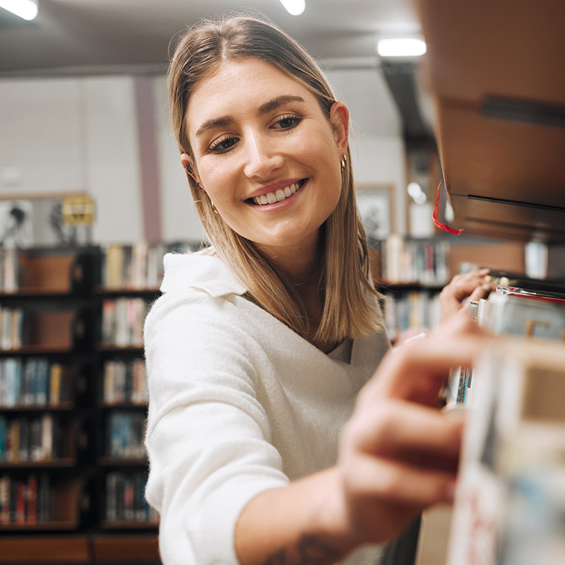 Smiling young woman browsing books in a library, showcasing Trillium Estates amenities with nearby public services and community resources.